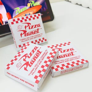 May include: Three red and white checkered pizza boxes with the words "Pizza Planet" and "Two Infinity and Beyond" printed on them. The boxes are for a birthday party and say "Thank you for coming to Allan's 2nd Birthday."