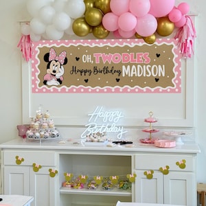 May include: A party scene with a banner that reads "Oh, Twodles Happy Birthday Madison" featuring Minnie Mouse. Balloons in white, gold, and pink are above the banner. Cupcakes, a neon sign, and decorations are on a white cabinet.