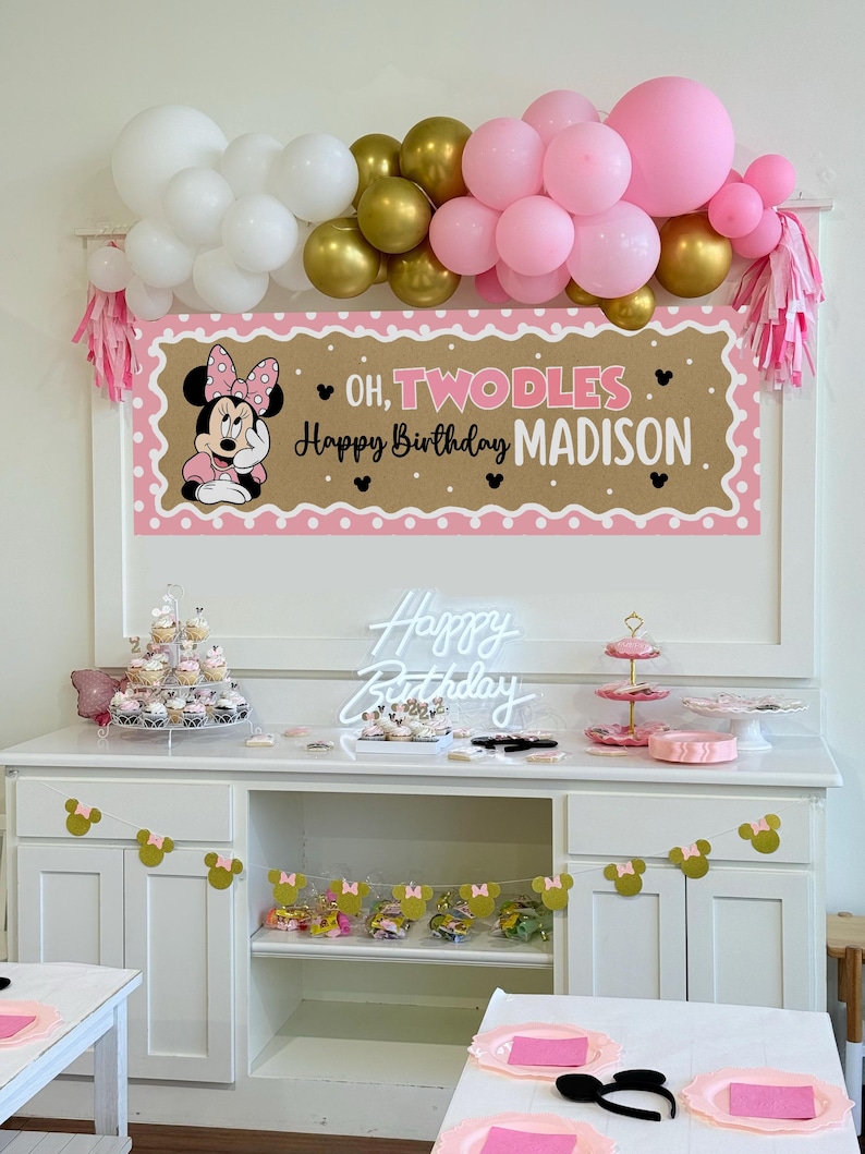 May include: A party scene with a banner that reads "Oh, Twodles Happy Birthday Madison" featuring Minnie Mouse. Balloons in white, gold, and pink are above the banner. Cupcakes, a neon sign, and decorations are on a white cabinet.