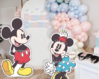 May include: Mickey Mouse and Minnie Mouse cardboard standups. Mickey is wearing red shorts and yellow shoes. Minnie is wearing a blue and white polka dot dress and yellow shoes. The standups are in front of a white backdrop with the words "Happiest Birthday" written on it. There are balloons in the background.