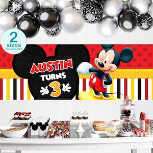 May include: A party banner featuring Mickey Mouse with the text "Austin Turns 3". The banner has a red background with white polka dots, yellow and black stripes, and a black Mickey Mouse head. The image also includes balloons and a table with desserts.