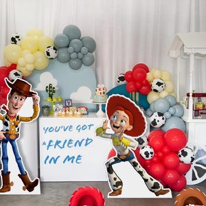 May include: A Toy Story themed party with a white candy cart, balloons, and cardboard cutouts of Woody and Jessie. The cart has the text "Andy's Candy Shack" on the side. The cardboard cutouts have the text "You've got a friend in me".