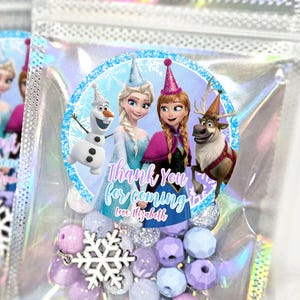 Könnte beinhalten: Eine transparente, schillernde Zellophantüte gefüllt mit Perlen und einem Schneeflocken-Anhänger. Die Tüte hat einen runden Aufkleber mit Figuren aus dem Film Frozen und dem Text "Thank You for coming".