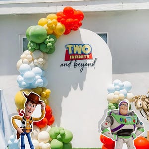 May include: A white backdrop with a balloon arch in shades of green, yellow, red, and blue. The backdrop has a sign that reads "TWO INFINITY and beyond". There are two cardboard cutouts of characters from the movie Toy Story: Woody and Buzz Lightyear.