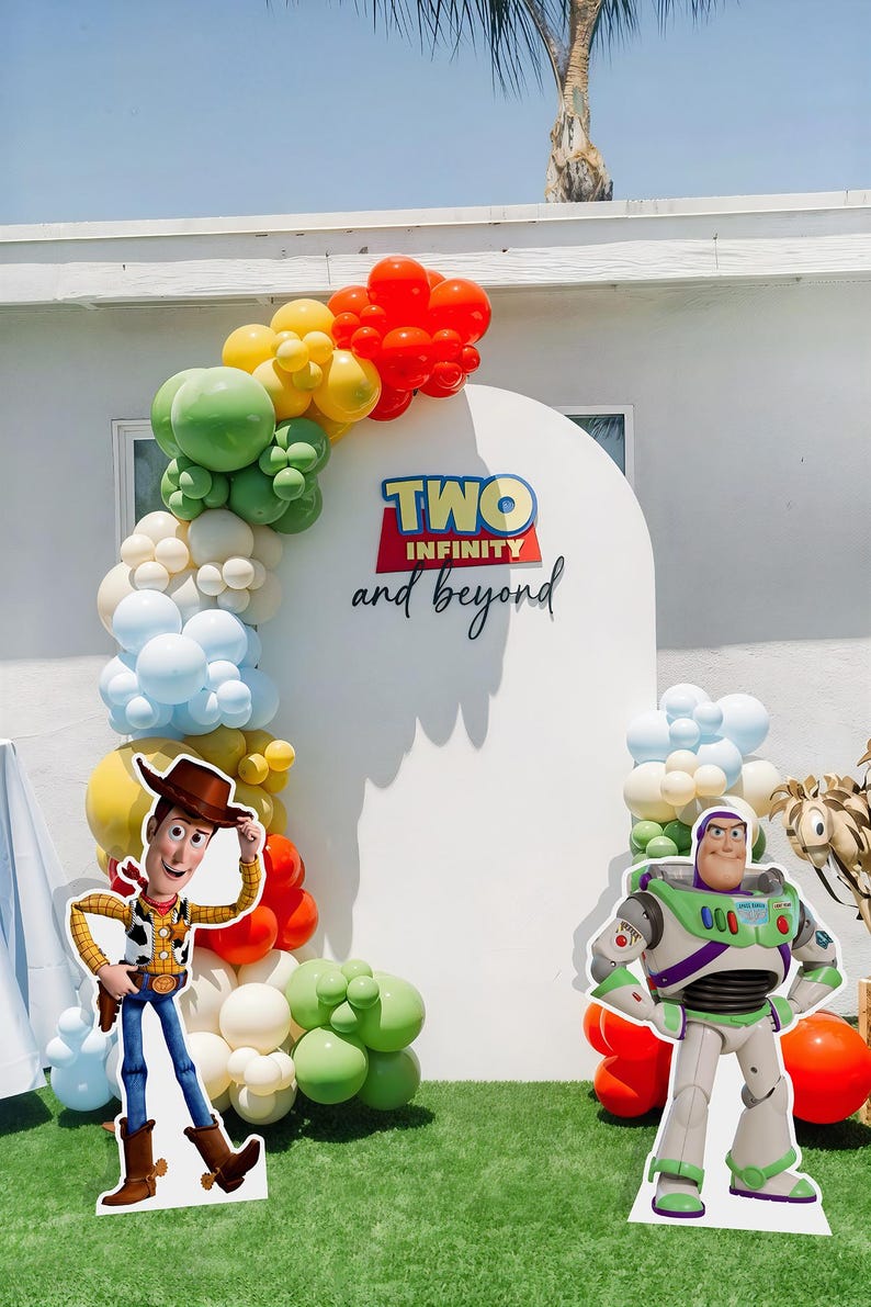 May include: A white backdrop with a balloon arch in shades of green, yellow, red, and blue. The backdrop has a sign that reads "TWO INFINITY and beyond". There are two cardboard cutouts of characters from the movie Toy Story: Woody and Buzz Lightyear.