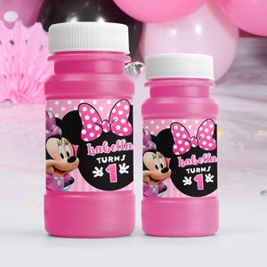 Peut inclure: Deux bouteilles de bulles en plastique rose avec des étiquettes sur le thème de Minnie Mouse. Les étiquettes représentent Minnie Mouse avec un nœud rose et des pois. Le texte sur les étiquettes indique "Isabella a 1 an".