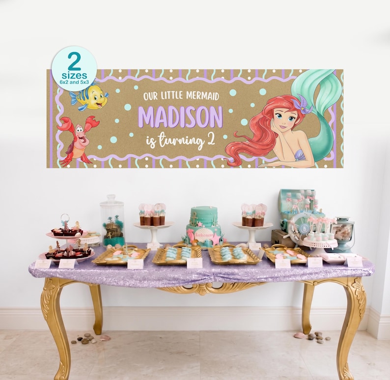 K&ouml;nnte beinhalten: Ein Banner mit dem Text "Our Little Mermaid Madison is turning 2" und Illustrationen zum Thema Meerjungfrau. Das Banner ist braun mit lila und wei&szlig;en Akzenten. Ein Tisch ist mit Desserts und Dekorationen gedeckt.