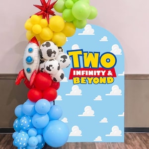 May include: A balloon arch featuring lime green, yellow, red, and light blue balloons, including a silver rocket ship balloon and white balloons with black cow spots. An arched backdrop displays a blue sky with white clouds and the text TWO INFINITY & BEYOND.