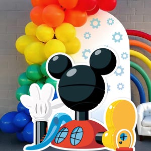 Op de afbeelding: Een Mickey Mouse-thema feestdecoratie met een grote Mickey Mouse-personage uitknippen, een regenboogballonboog en een witte achtergrond met blauwe tandwielen.