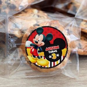 Op de afbeelding: Een chocolate chip cookie in een doorzichtige plastic zak met een Mickey Mouse sticker. De sticker heeft een rode achtergrond met Mickey Mouse en de tekst "Austin Turns 3". De cookie is rond en heeft een verscheidenheid aan chocolate chips.