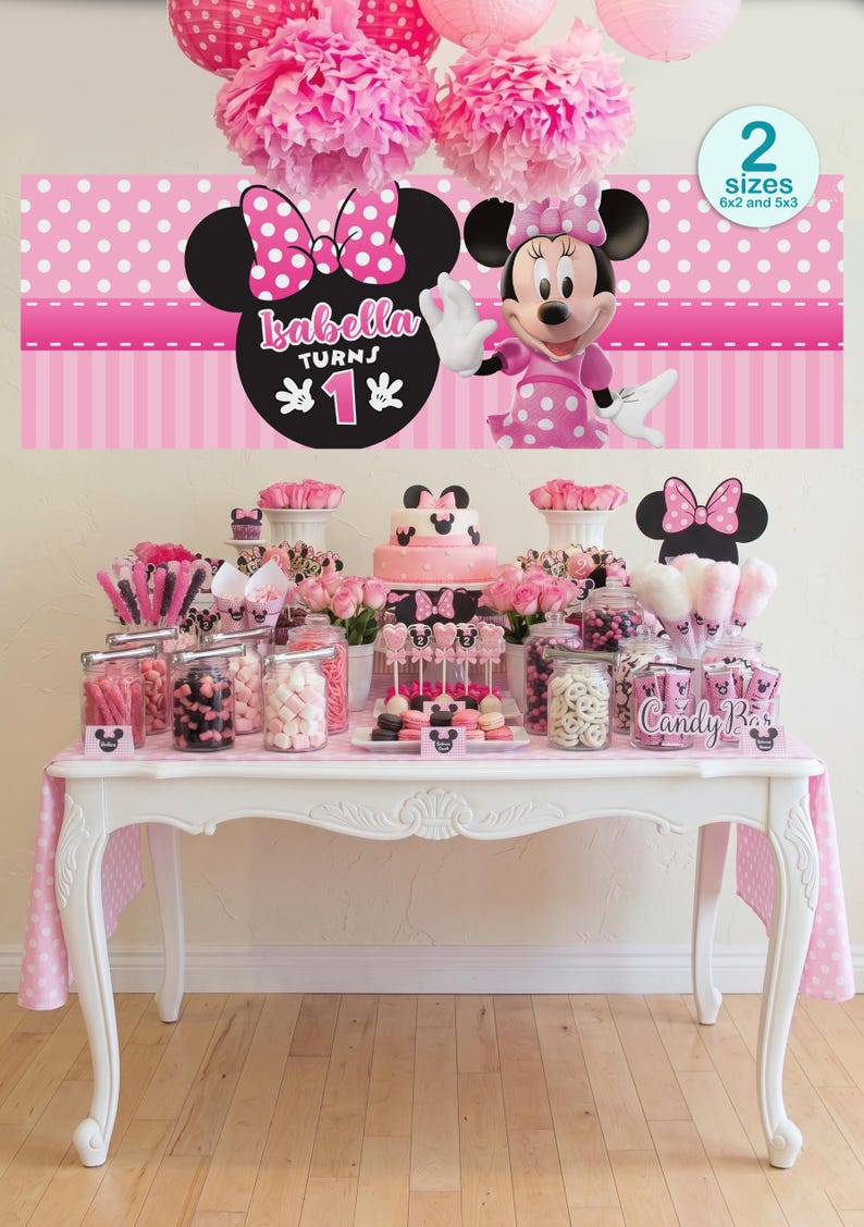 Op de afbeelding: Een roze Minnie Mouse themafeestopstelling. De tafel is versierd met een roze tafelkleed, lekkernijen en een taart. Een banner met Minnie Mouse en de tekst "Isabella Turns 1" is boven de tafel te zien. Roze pompons en papieren lantaarns hangen aan het plafond.