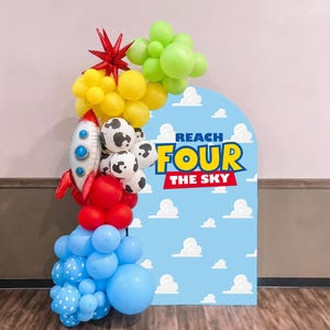 Puede incluir: Un arco de globos amarillos, verde lima, rojos y azules, incluyendo globos con estampado de vaca y un globo de aluminio con forma de cohete. Junto a él, un panel arqueado con cielo azul, nubes blancas y el texto REACH FOUR THE SKY.