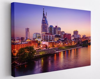 Może przedstawiać: Panoramiczny widok na panoramę Nashville o zmierzchu, z charakterystycznym budynkiem AT&T z jego charakterystyczną iglicą. Miasto jest oświetlone latarniami ulicznymi, które odbijają się w spokojnych wodach rzeki Cumberland.