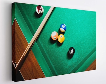 May include: A green felt pool table with a wooden side rail. There are six billiard balls on the table, including a red ball, a blue ball, a yellow ball, a white ball, a black ball, and a ball with the number 13 on it. A wooden pool cue is lying on the table.