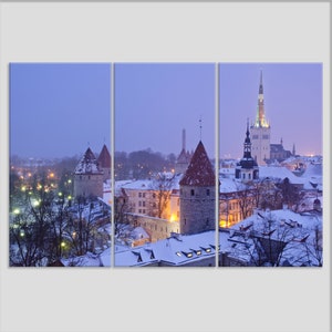May include: A panoramic view of a snow-covered cityscape at dusk. The scene features several medieval towers and a tall church steeple with a pointed roof. The buildings are illuminated by streetlights, creating a warm glow against the purple sky.
