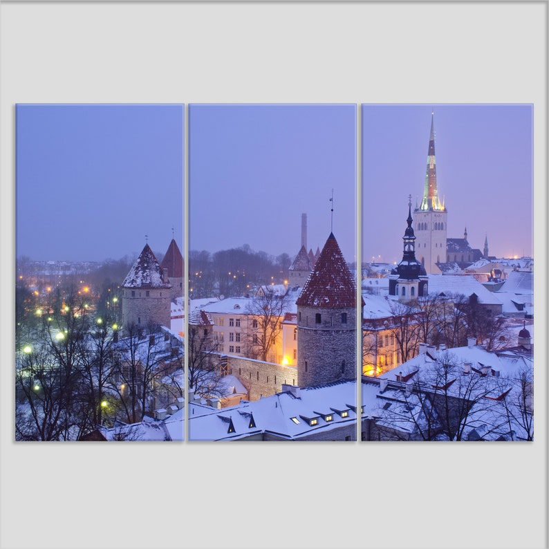 May include: A panoramic view of a snow-covered cityscape at dusk. The scene features several medieval towers and a tall church steeple with a pointed roof. The buildings are illuminated by streetlights, creating a warm glow against the purple sky.