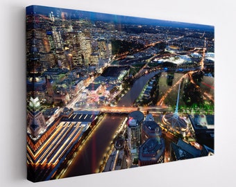 May include: An aerial night view of a city with a river running through it. The city is lit up with streetlights and buildings. There is a large stadium in the distance.