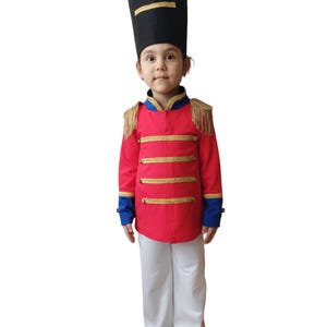 May include: A child wearing a red and gold toy soldier costume with a black hat. The costume has gold trim and blue accents on the sleeves.