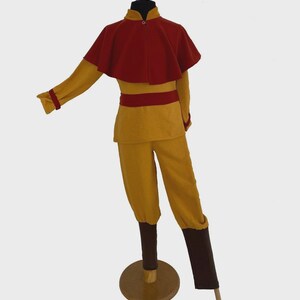 Puede incluir: Un conjunto de disfraz compuesto por una camiseta de manga larga color amarillo mostaza con una capa corta roja, una faja roja a juego, pantalones amarillo mostaza y cubrepiernas de color marr&oacute;n oscuro sobre un maniqu&iacute;.
