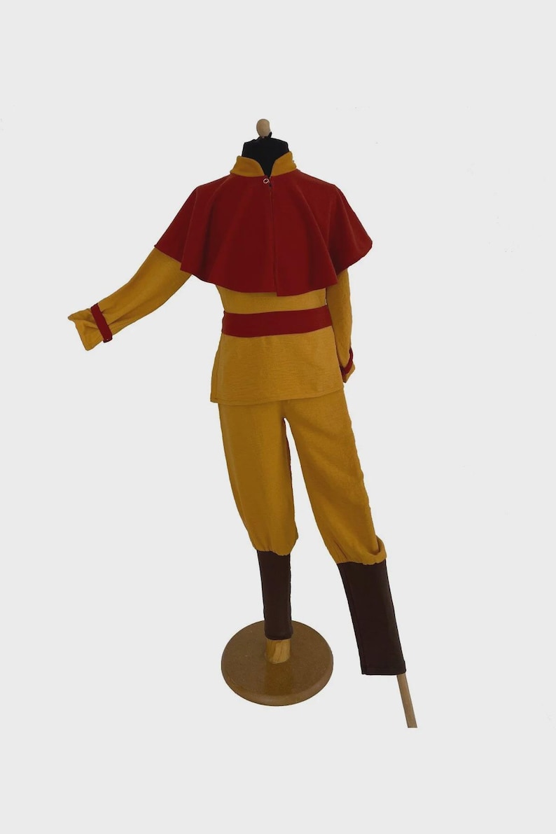 Puede incluir: Un conjunto de disfraz compuesto por una camiseta de manga larga color amarillo mostaza con una capa corta roja, una faja roja a juego, pantalones amarillo mostaza y cubrepiernas de color marr&oacute;n oscuro sobre un maniqu&iacute;.