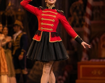 May include: A red and black military-style costume featuring a structured jacket with gold braided trim, epaulets, and a high collar. The outfit includes a black tulle skirt, a tall black cylindrical hat with a gold V-shaped detail, and black boots.