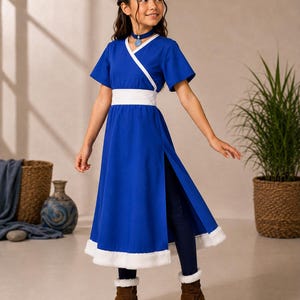 May include: A vibrant blue wrap-style dress featuring a white V-neck trim, a wide white waistband, and a white faux fur hem. The dress has short sleeves and a high side slit, worn over dark blue leggings and brown suede boots with white fur cuffs.
