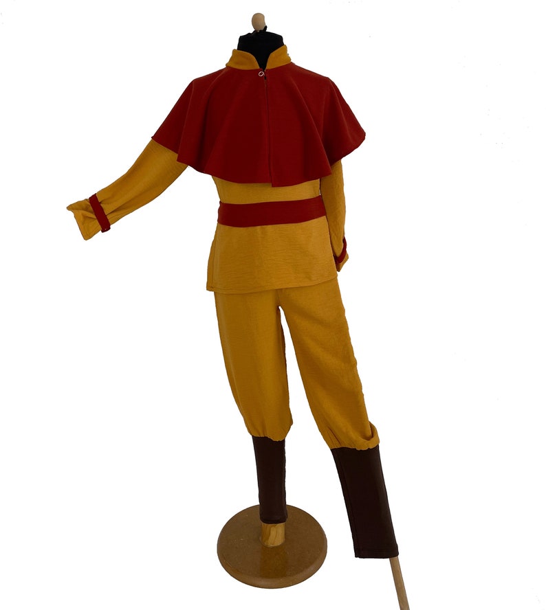 Puede incluir: Un conjunto de t&uacute;nica de manga larga y pantalones color amarillo mostaza que presenta una capa corta de color rojo oscuro unida al cuello, una faja roja y cubrepiernas de color marr&oacute;n oscuro. El atuendo se exhibe sobre un maniqu&iacute;.