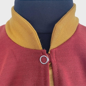 Puede incluir: Una chaqueta granate con cuello acanalado de color amarillo mostaza y un cierre de bot&oacute;n a presi&oacute;n de metal plateado en el escote. La prenda se muestra sobre un maniqu&iacute; azul oscuro, resaltando el contraste entre los colores.