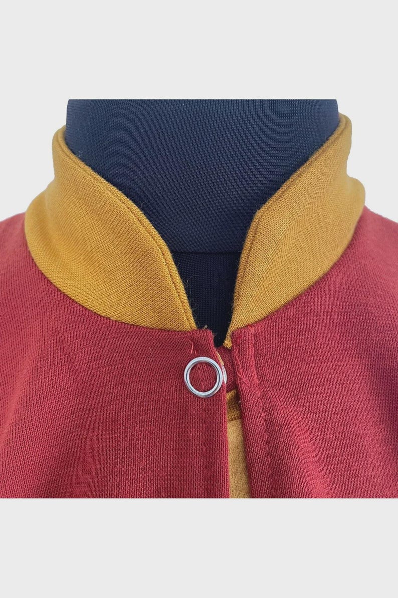 Puede incluir: Una chaqueta granate con cuello acanalado de color amarillo mostaza y un cierre de bot&oacute;n a presi&oacute;n de metal plateado en el escote. La prenda se muestra sobre un maniqu&iacute; azul oscuro, resaltando el contraste entre los colores.