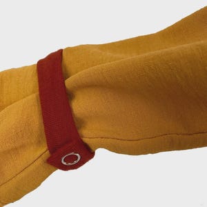 Puede incluir: Una prenda de manga larga color amarillo mostaza con una tira de tela de color rojo oscuro en contraste en el pu&ntilde;o. La tira se asegura con un bot&oacute;n de presi&oacute;n de metal plateado, proporcionando un detalle de cierre funcional en la manga.
