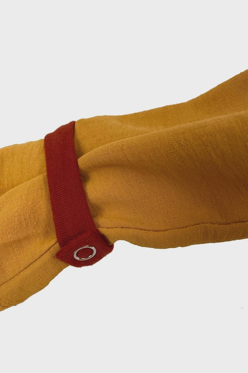 Puede incluir: Una prenda de manga larga color amarillo mostaza con una tira de tela de color rojo oscuro en contraste en el pu&ntilde;o. La tira se asegura con un bot&oacute;n de presi&oacute;n de metal plateado, proporcionando un detalle de cierre funcional en la manga.