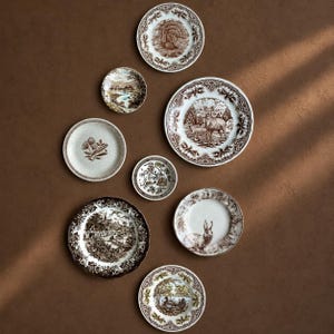 May include: A collection of eight ceramic plates featuring brown transferware patterns. The designs include a turkey, a deer in a forest, a rabbit, a landscape with a village, a floral motif, and various nature scenes with intricate decorative borders.