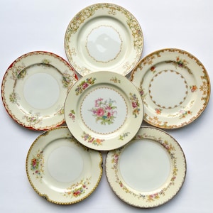 May include: A collection of five round ceramic plates arranged in a cluster. Each plate features a cream-coloured base with distinct decorative borders, including floral patterns, gold-toned filigree, and scalloped edges in red and brown hues.