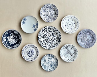 May include: A collection of eight round ceramic plates arranged in a cluster. Each plate features a distinct blue and white pattern, ranging from dense floral and botanical illustrations to intricate geometric borders and solid blue backgrounds.