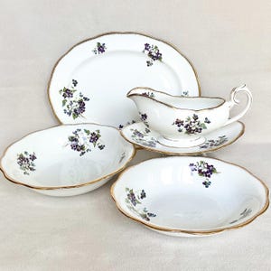 May include: A white porcelain dinnerware set featuring a floral design with purple and green accents. The set includes a platter, gravy boat with saucer, and two oval serving bowls. Each piece has a gold rim.