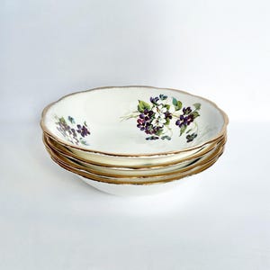 May include: Four white ceramic bowls with a scalloped edge and gold trim. Each bowl has a floral design with purple and white flowers and green leaves. The bowls are stacked. The bowls are approximately 20 cm in diameter.