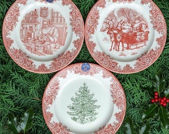 May include: Three decorative Christmas plates with a red and white floral border. The plates feature festive scenes: Santa in his workshop, Santa in a sleigh with reindeer, and a Christmas tree. The plates are set on a bed of green foliage.