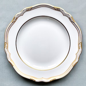 May include: A white porcelain plate with a scalloped edge and gold trim. The plate features a gold band around the rim and a decorative gold pattern along the edge. The plate is a classic piece of dinnerware.
