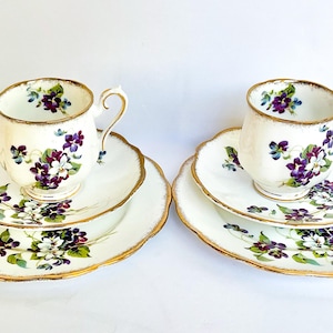May include: A pair of teacups and saucers. The white porcelain has a floral pattern of purple and white violets, with gold edging. The cups sit on matching saucers with a scalloped edge, adding a classic touch.