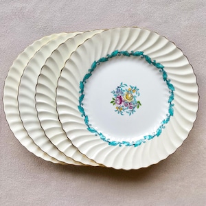 May include: Four cream-coloured plates with a gold rim and a ruffled edge design. Each plate has a teal floral border and a central floral bouquet in pink, yellow, and blue. The plates are stacked. The plates are suitable for serving food.