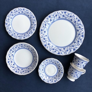 May include: A collection of white porcelain tableware with a blue floral design. The set includes plates, saucers, and teacups. The plates have scalloped edges, and the teacups have a pedestal base.