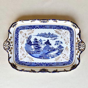 May include: Rectangular serving tray with a blue and white Asian-inspired design. The tray features a detailed landscape scene with pagodas, trees, and a bridge. Gold accents adorn the edges and handles, adding a touch of elegance to this decorative piece.