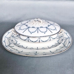 Peut inclure: Un plat de service en porcelaine blanche avec une bordure bleue et dorée. Le plat a un couvercle et un plat ovale assorti. Le plat est décoré d'un motif répétitif de guirlandes.