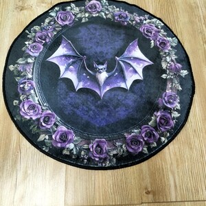 Puede incluir: Una alfombra redonda de color negro y morado con un murci&eacute;lago en el centro y un borde de rosas moradas. El murci&eacute;lago est&aacute; volando con las alas extendidas y tiene la cara blanca. Las rosas est&aacute;n dispuestas en un c&iacute;rculo alrededor del murci&eacute;lago.