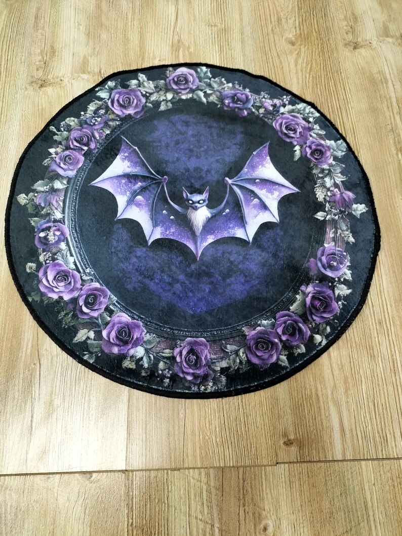 Puede incluir: Una alfombra redonda de color negro y morado con un murci&eacute;lago en el centro y un borde de rosas moradas. El murci&eacute;lago est&aacute; volando con las alas extendidas y tiene la cara blanca. Las rosas est&aacute;n dispuestas en un c&iacute;rculo alrededor del murci&eacute;lago.