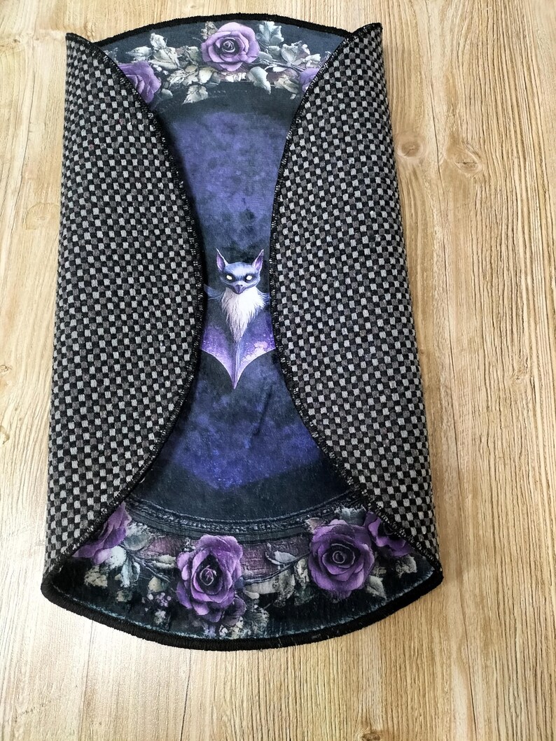 Puede incluir: Una alfombra redonda de color negro y morado con un dise&ntilde;o de murci&eacute;lago y rosas. La alfombra tiene un patr&oacute;n a cuadros en los bordes.