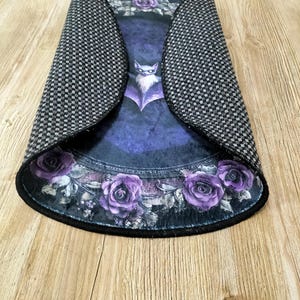 Puede incluir: Una alfombra redonda de color morado y negro con un dise&ntilde;o de murci&eacute;lago y rosas. La alfombra tiene un borde negro y un respaldo antideslizante.