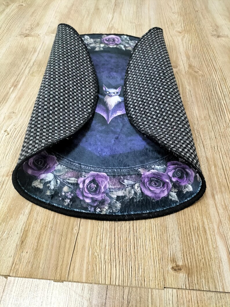 Puede incluir: Una alfombra redonda de color morado y negro con un dise&ntilde;o de murci&eacute;lago y rosas. La alfombra tiene un borde negro y un respaldo antideslizante.