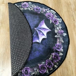 Puede incluir: Una alfombra redonda de color morado y negro con un dise&ntilde;o g&oacute;tico que presenta un ala de drag&oacute;n y una corona de rosas moradas. La alfombra tiene un respaldo antideslizante.