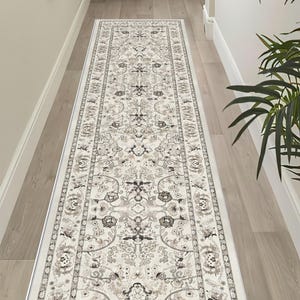 May include: A long rectangular runner rug featuring a cream-colored base with an intricate floral and vine pattern in shades of charcoal gray, muted brown, and soft beige. The design includes a decorative border with repeating geometric motifs.
